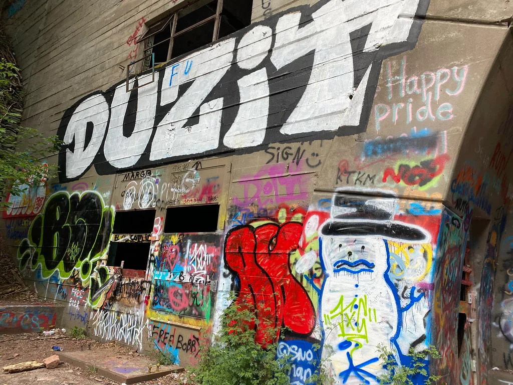 Façade industrielle abandonnée couverte de graffitis, avec baies vides et portes percées.