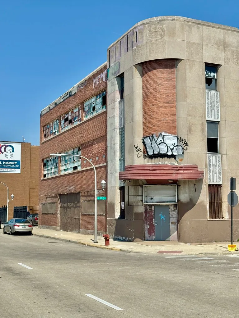 Old brick building with shattered windows and a boarded entrance.