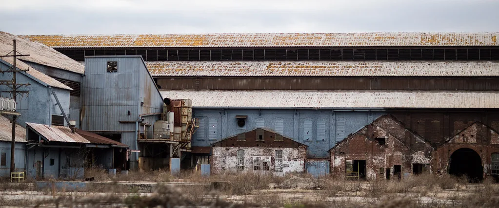 Old brick and metal industrial halls beside overgrown ground.