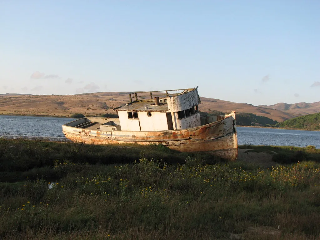 Old white wooden boat stranded beside the water with a peeling hull and deteriorated cabin.