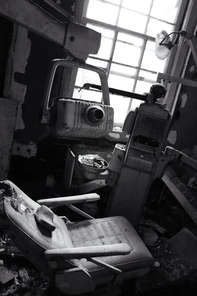 Abandoned dental room with a torn chair and old equipment in front of a large window, in black and white.
