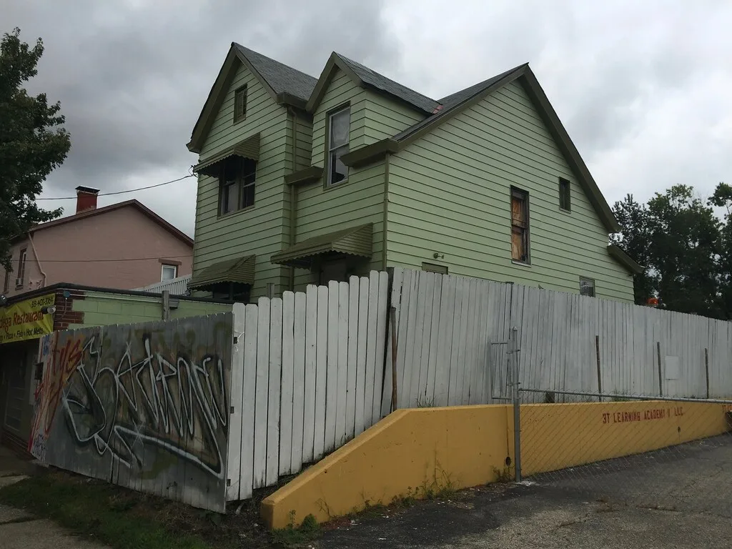 Maison abandonnée à bardage vert et pignons multiples, aux vitres brisées ou condamnées derrière une palissade blanche graffitée.