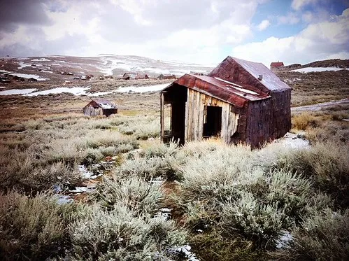 Small derelict wooden cabin with a rusty roof amid scrub and patches of snow.