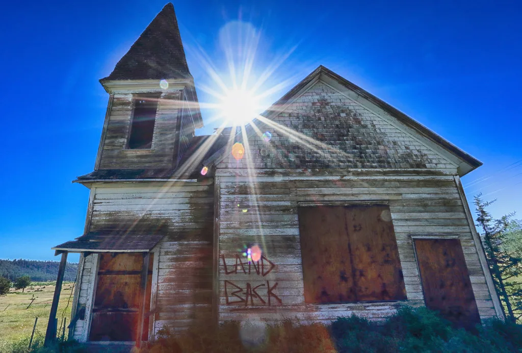 Abandoned church with peeling white siding, boarded openings and a dark bell tower beneath the sun.