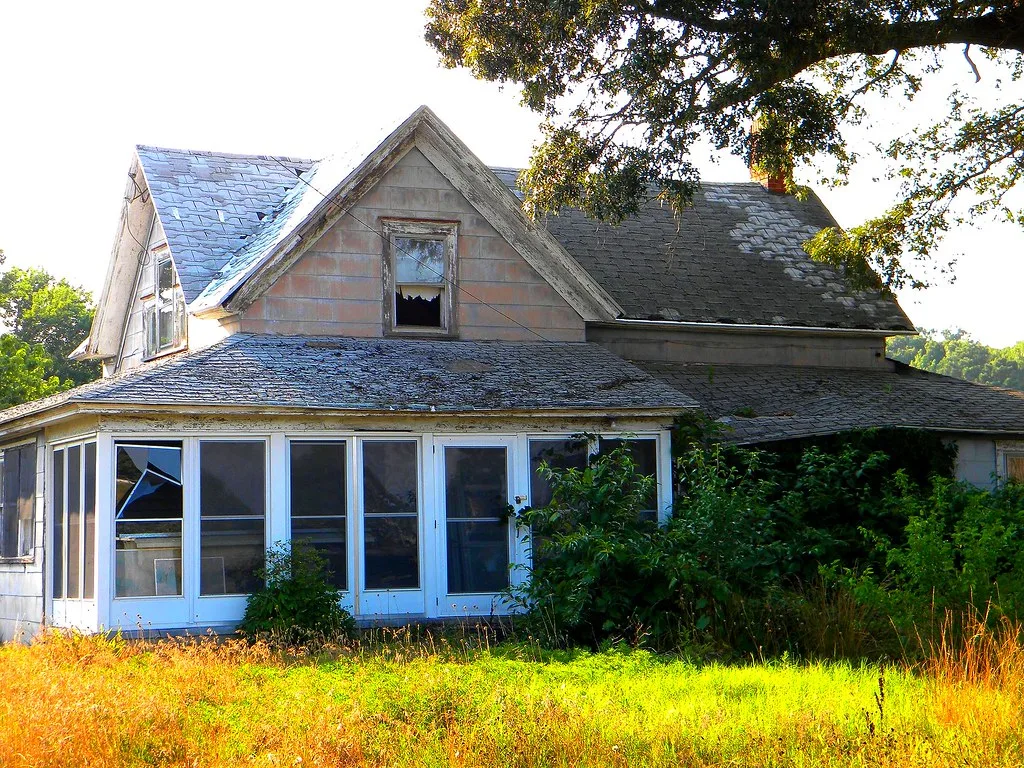 Old house with broken windows surrounded by tall grass and shrubs.