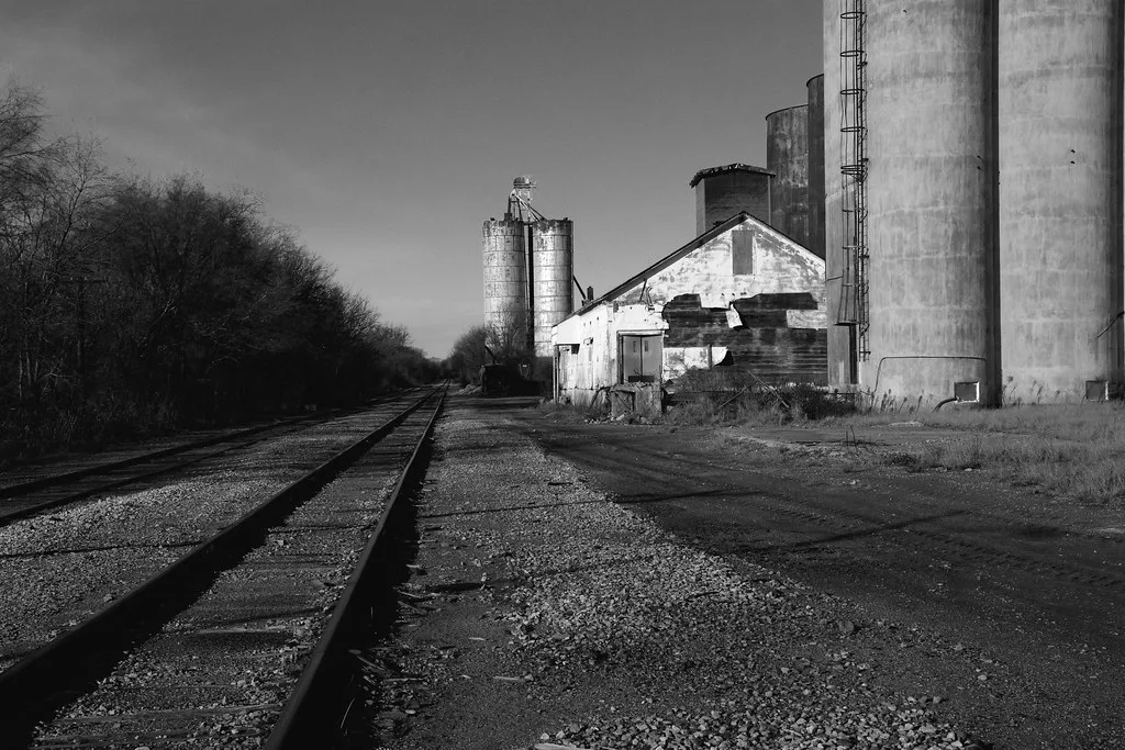 Abandoned old warehouse with torn white cladding beside silos and a railway track, in black and white.