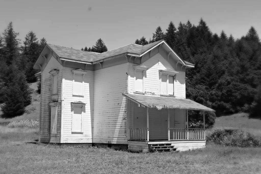 White house with a covered porch and all windows sealed in a meadow.