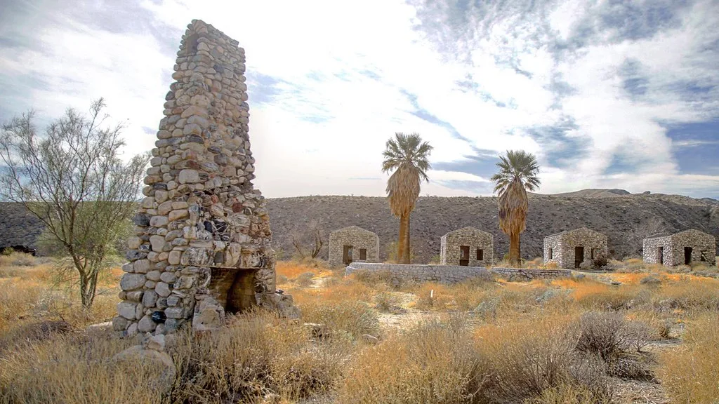 Old stone chimney in front of ruined houses and palm trees.