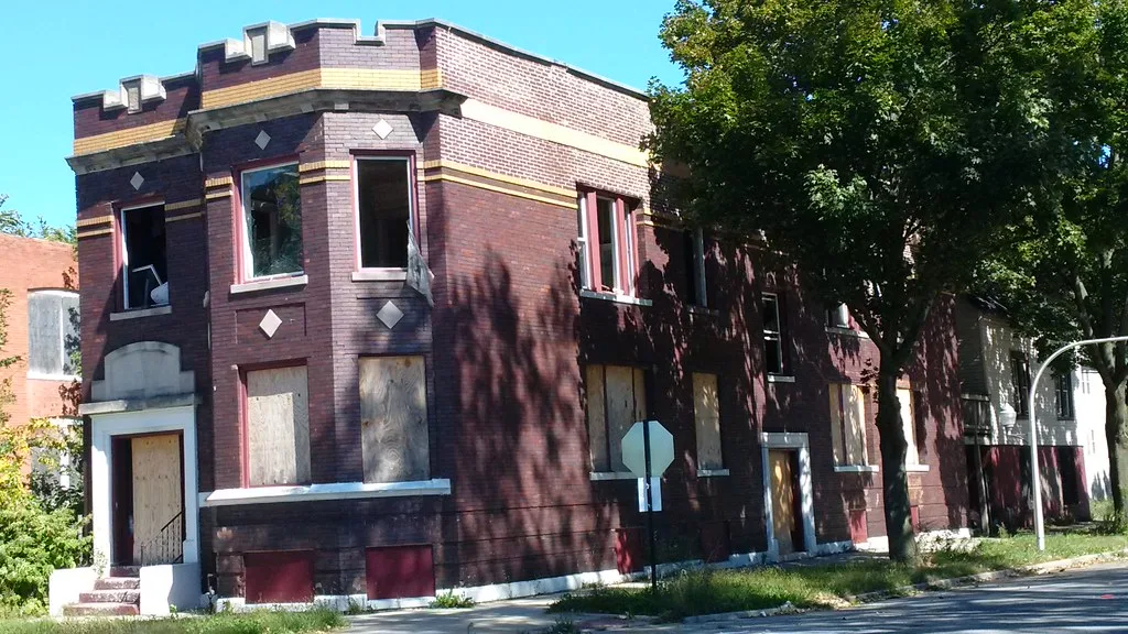 Dark brick corner building with broken and boarded windows.