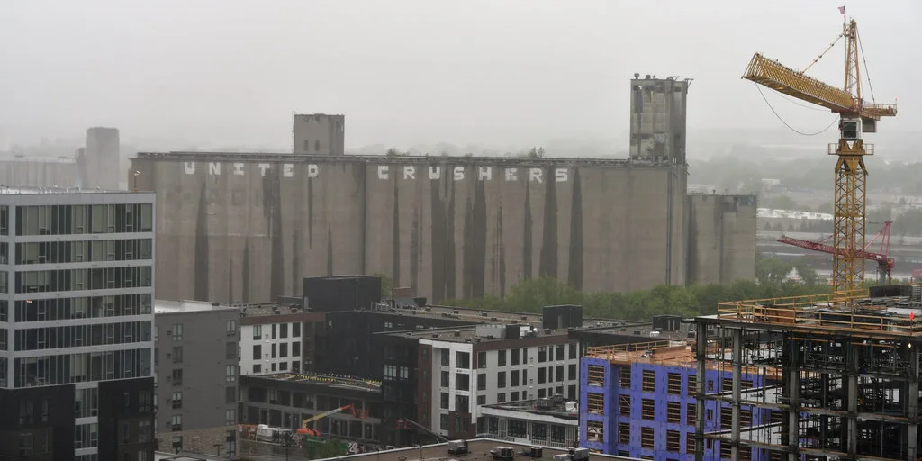 Large deteriorated concrete silo complex behind a neighborhood under construction beneath a gray sky.