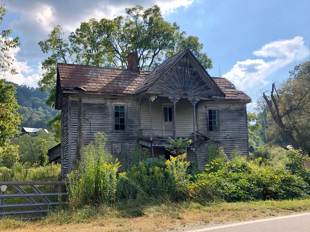 Maison de bois délabrée au milieu des hautes herbes.