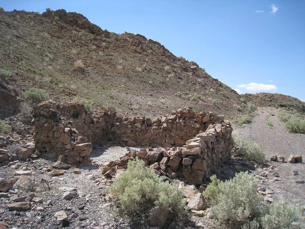 Ruins of a small roofless stone structure at the foot of a rocky slope, surrounded by grey shrubs.
