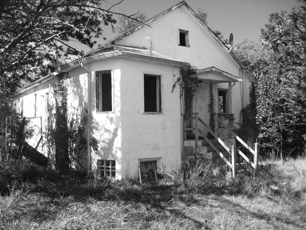 Abandoned white house with empty windows and an exterior wooden staircase.