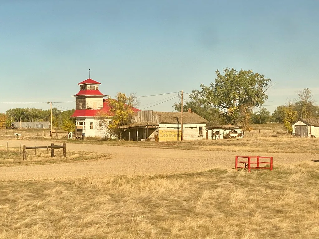 Old rural shop with a red tower and dilapidated outbuildings amid dry grass.