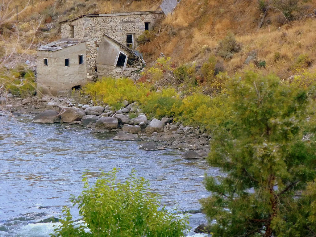 Abandoned stone and concrete buildings beside a river, with a collapsed annex on a grassy slope.
