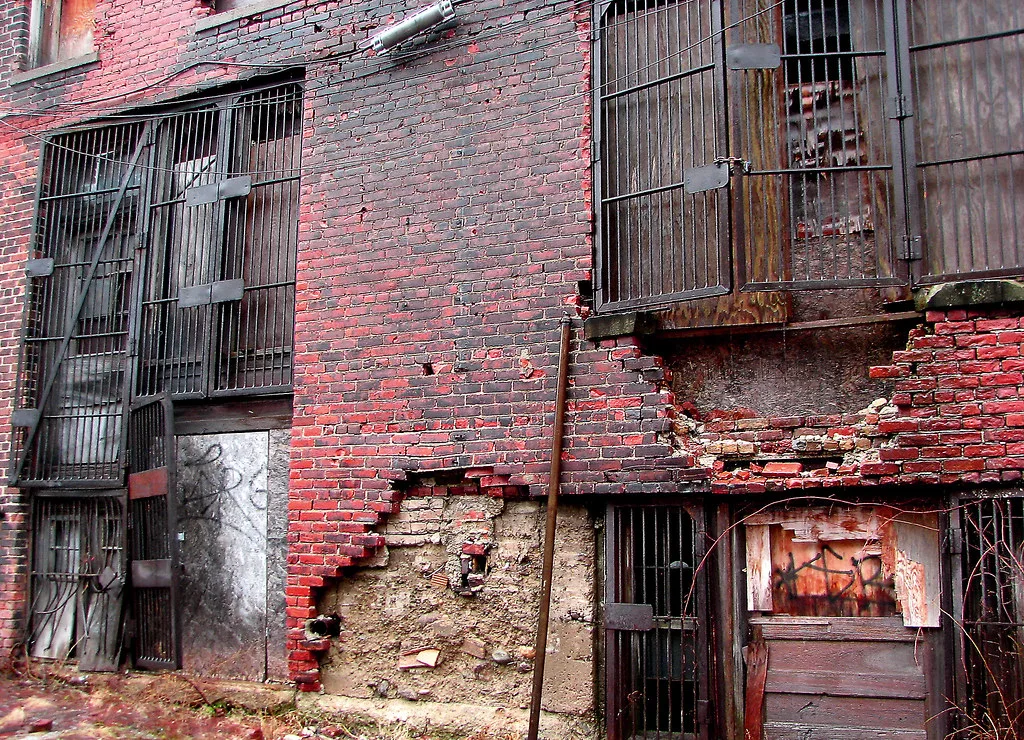 Deteriorated brick facade with boarded openings and rusty grilles.