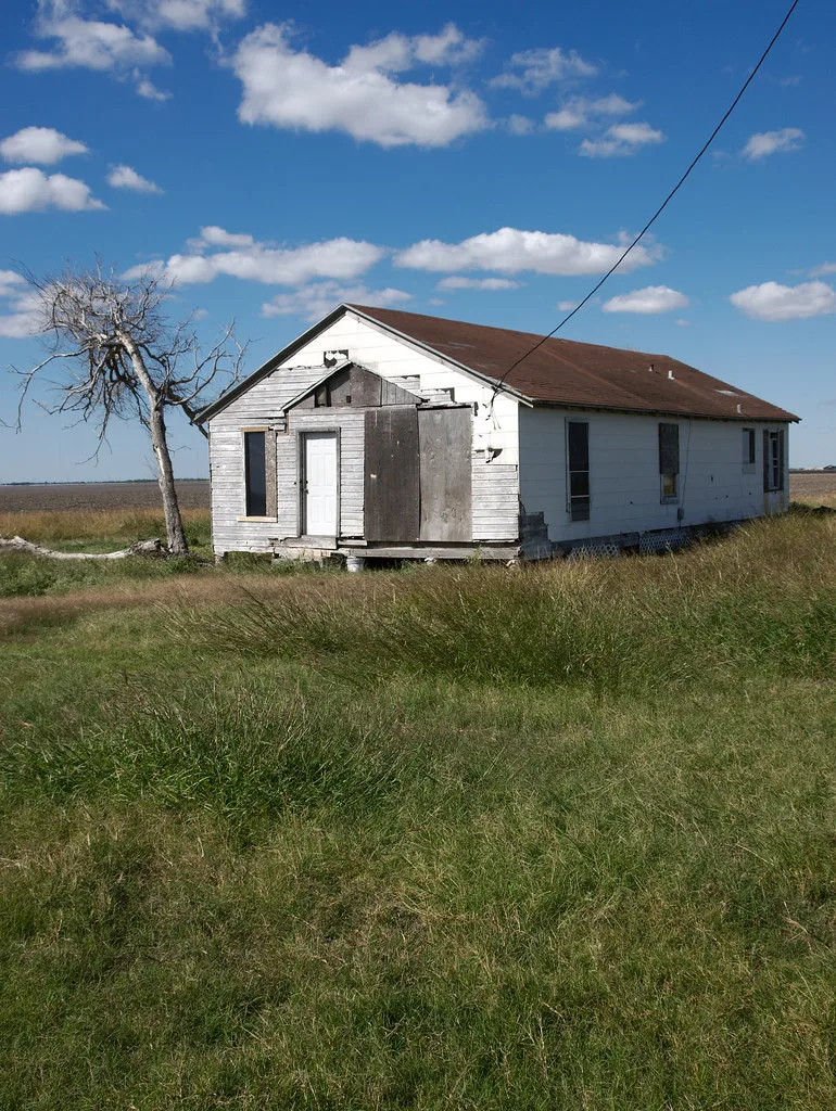 Small white house with boarded openings in tall grass.