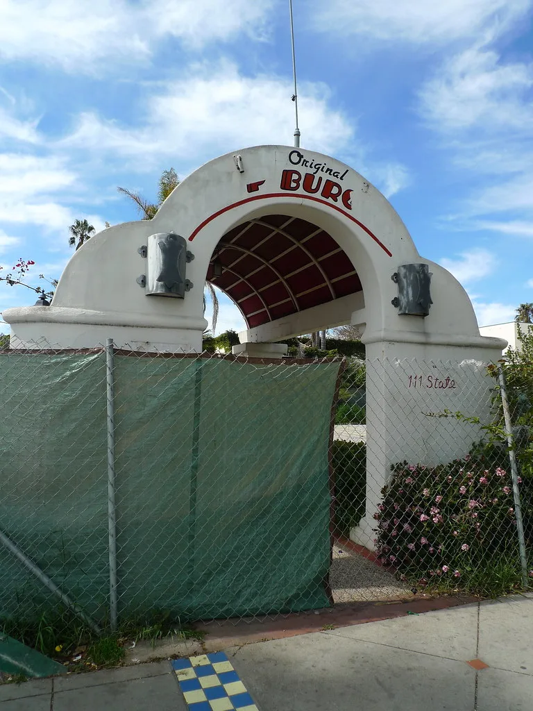 Old arched entrance behind a fence, with an incomplete sign and vegetation.
