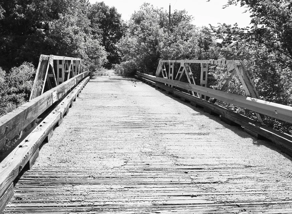Old metal bridge with a deteriorated wooden deck amid vegetation, in black and white.