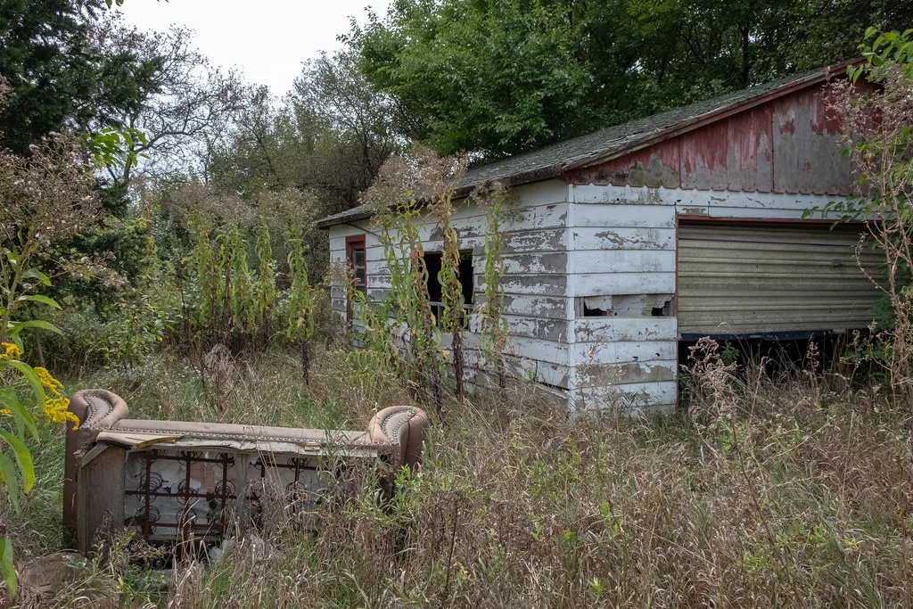 Old white and red garage overgrown with vegetation, with an overturned sofa in front.