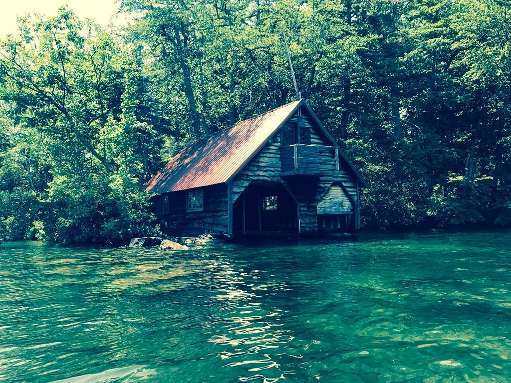 Ancien abri à bateaux en bois au bord d’une eau verte devant une forêt.