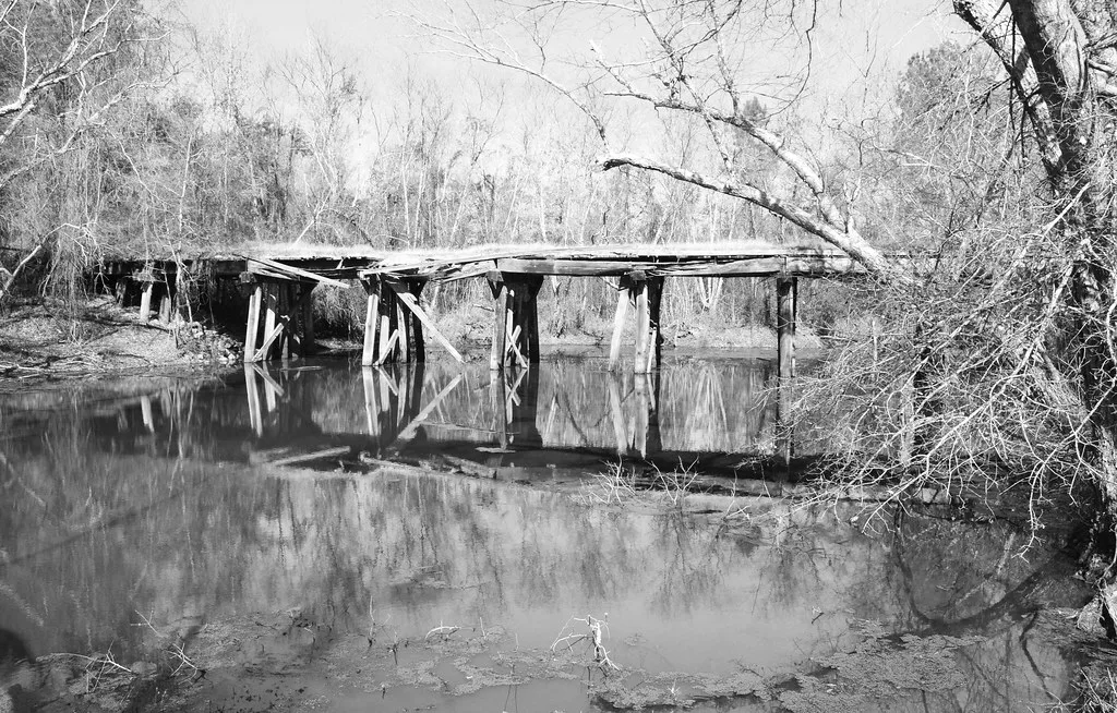 Old wooden bridge with a broken deck reflected in a river, in black and white.