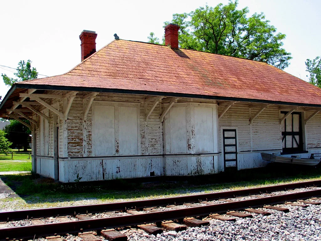 Old white wooden station with sealed windows and peeling paint beside railway tracks.