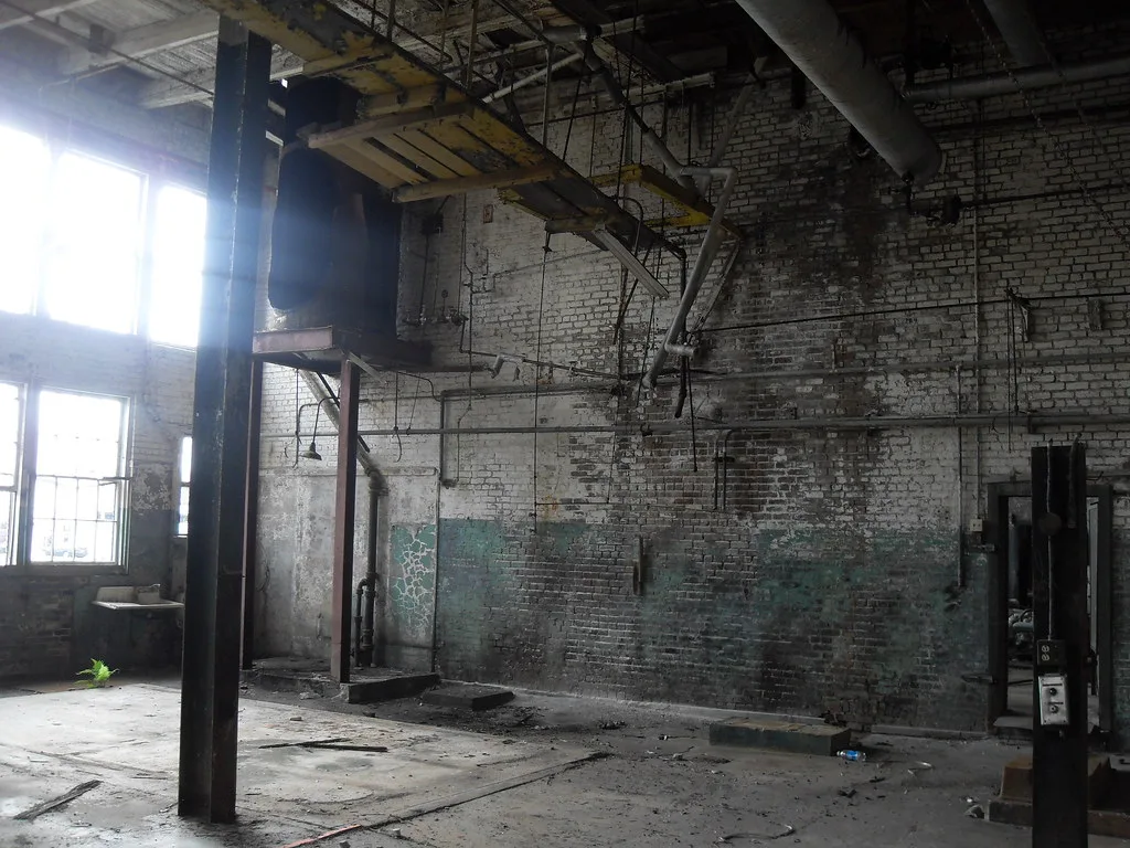 Abandoned factory room with flaking brick walls, hanging pipes and remnants of machinery.