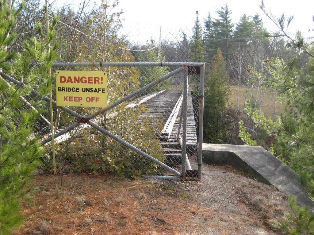 Old abandoned bridge with rails and sleepers overgrown by vegetation, closed by a gate carrying a danger warning.