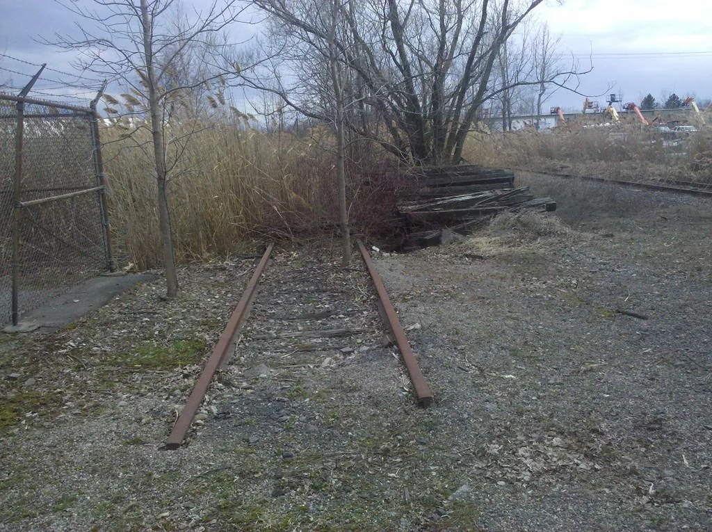 Old railway track interrupted by a tree beside a pile of sleepers and a fence.