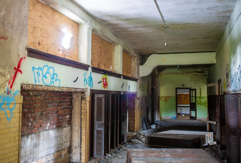 Abandoned corridor with boarded windows, graffitied walls and bricked-up openings, with removed metal doors and debris on the floor.