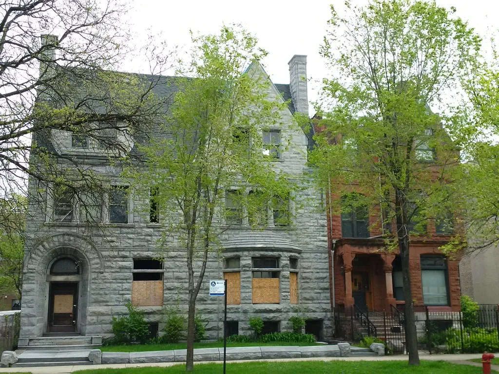 Large neglected gray-stone house with broken windows and boarded ground-floor openings behind trees.