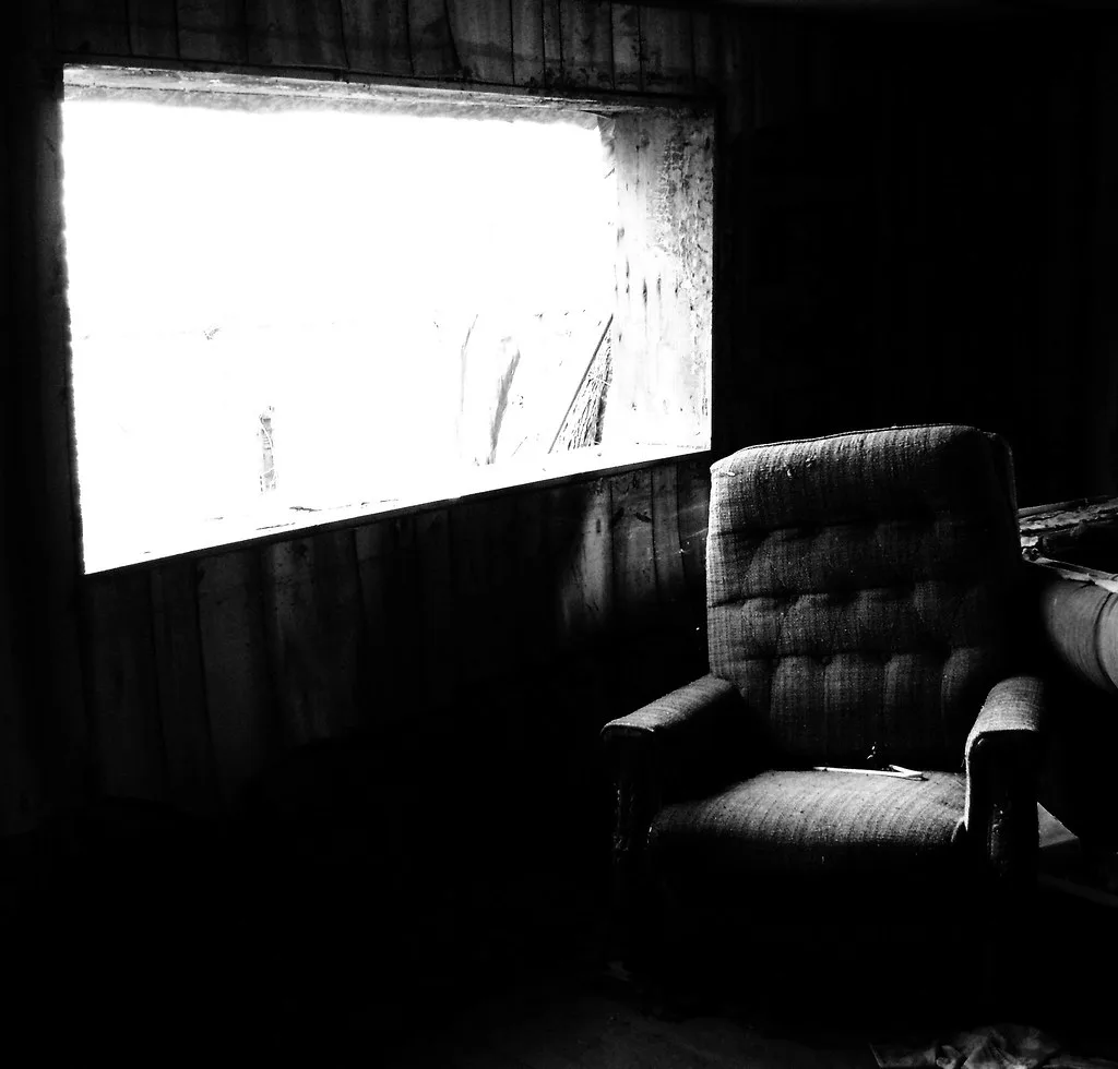Fauteuil dans une pièce dégradée éclairée par une grande ouverture, en noir et blanc.