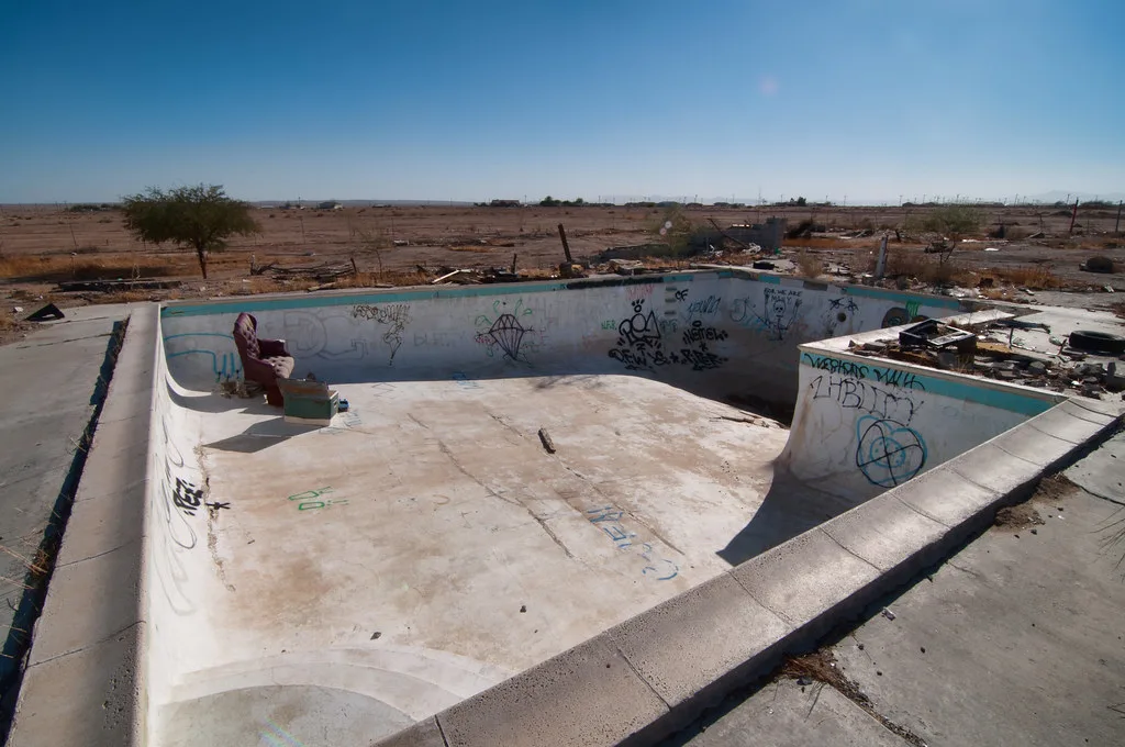 Piscine abandonnée vide et couverte de graffitis, avec un fauteuil au fond au milieu d’un terrain aride.
