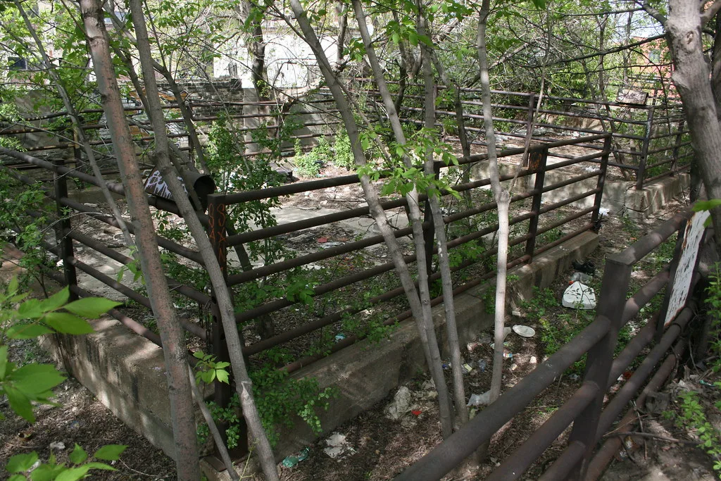 Anciens enclos aux barrières rouillées et soubassements de béton envahis par des arbres et des déchets.