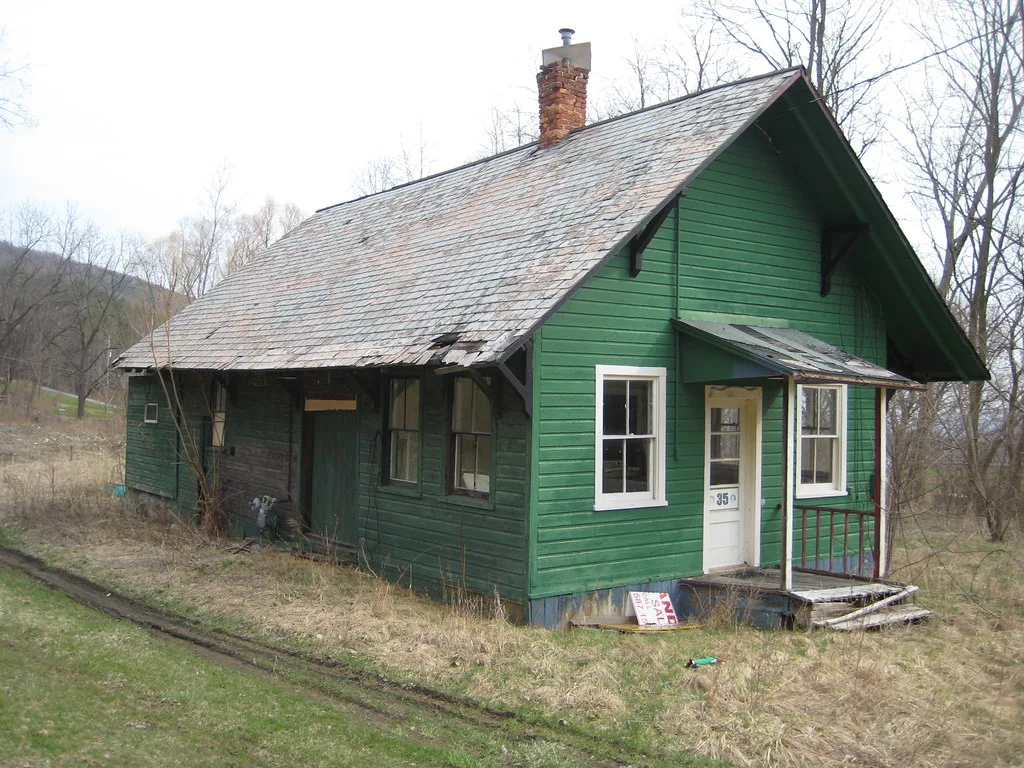 Small green wooden house with a damaged roof and boarded windows.