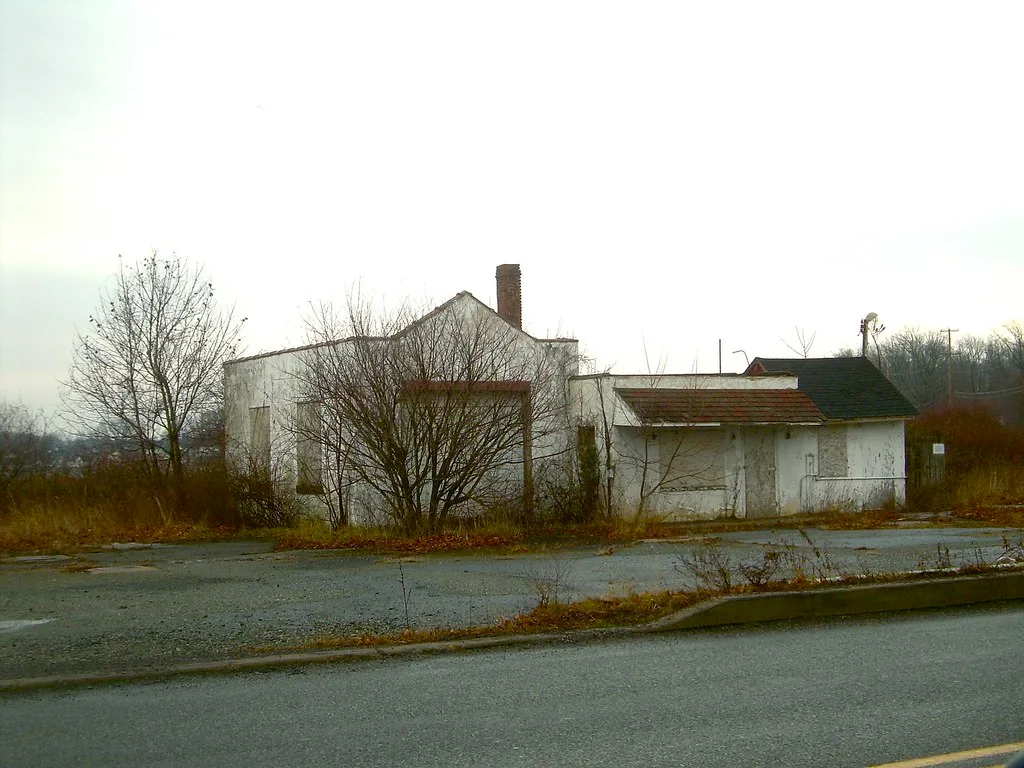 Old white station building with boarded openings and overgrown surroundings.