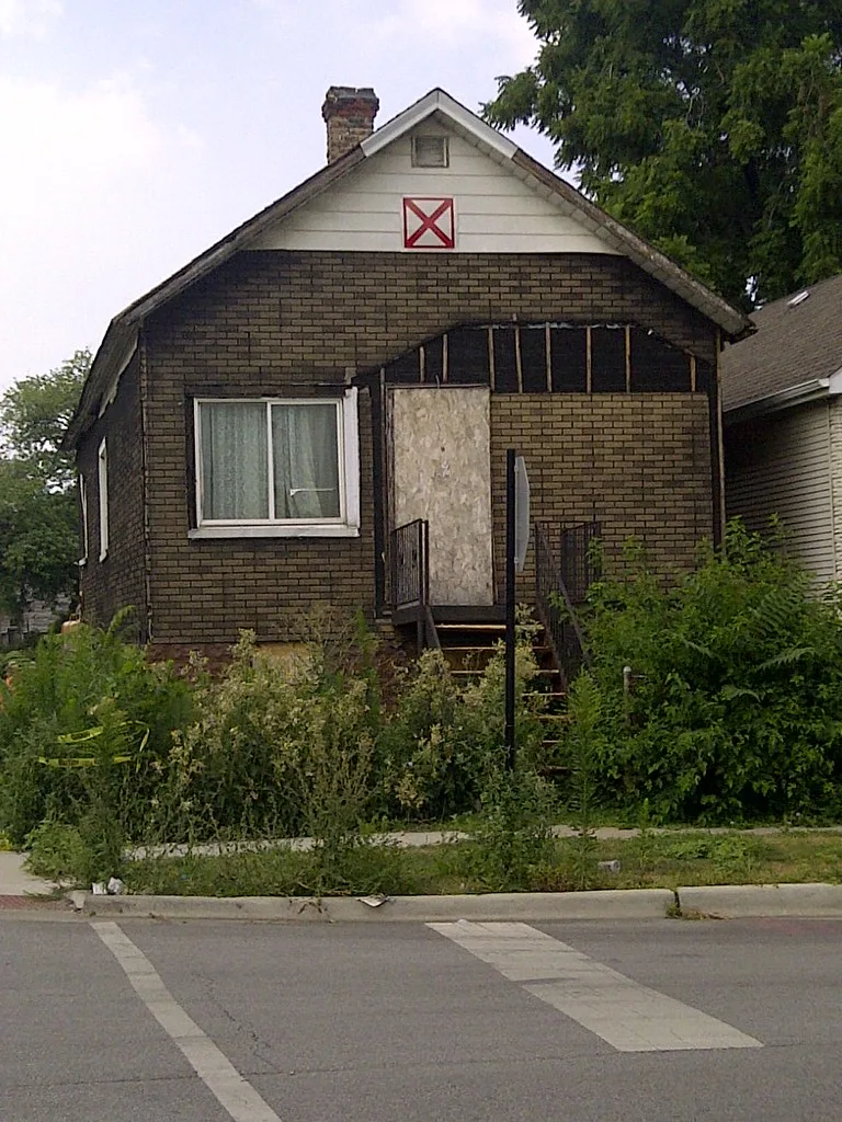 Small abandoned brick house with a boarded entrance and vegetation covering the steps.
