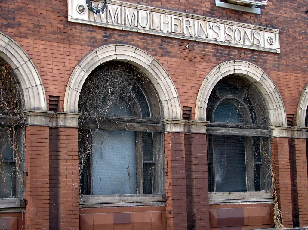 Brick facade with deteriorated arched windows overgrown with dry vines.