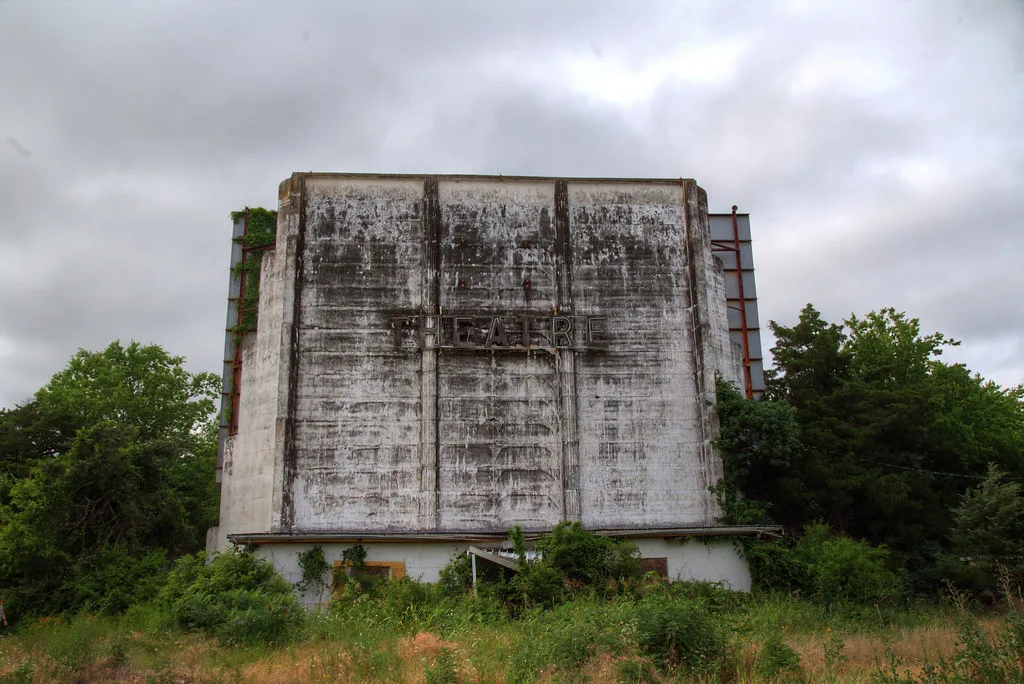 Ancien écran de cinéma abandonné aux panneaux noircis et bâtiment bas condamné, envahi par les arbustes et hautes herbes.
