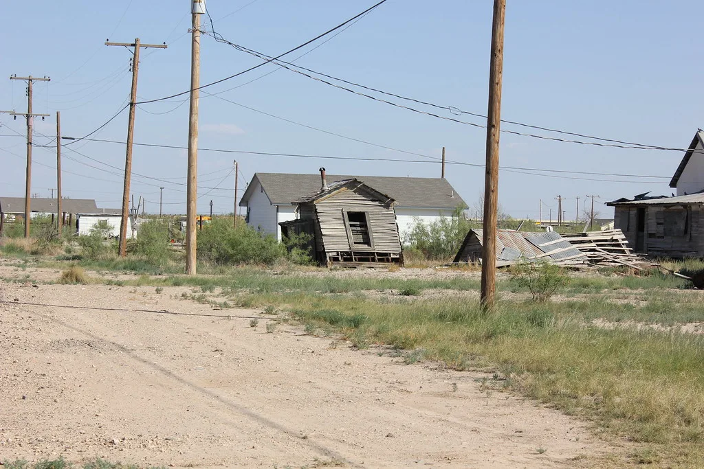 Small leaning abandoned wooden house with an empty doorway beside collapsed sheet metal and framing on grassy ground.