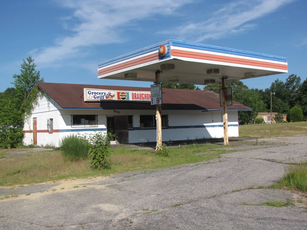 Ancienne station-service délaissée avec auvent sans pompes et boutique devant une aire envahie d’herbes.