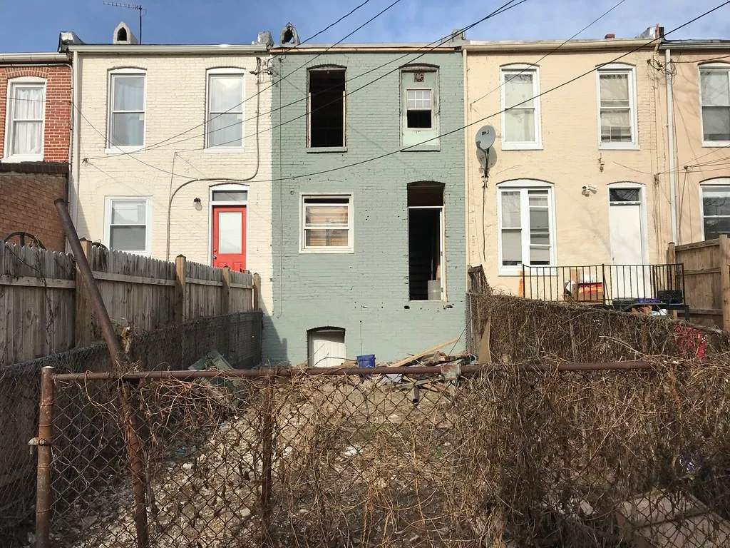 Arrière d’une maison abandonnée en brique vert pâle aux baies vides, derrière une cour encombrée de débris et un grillage rouillé.
