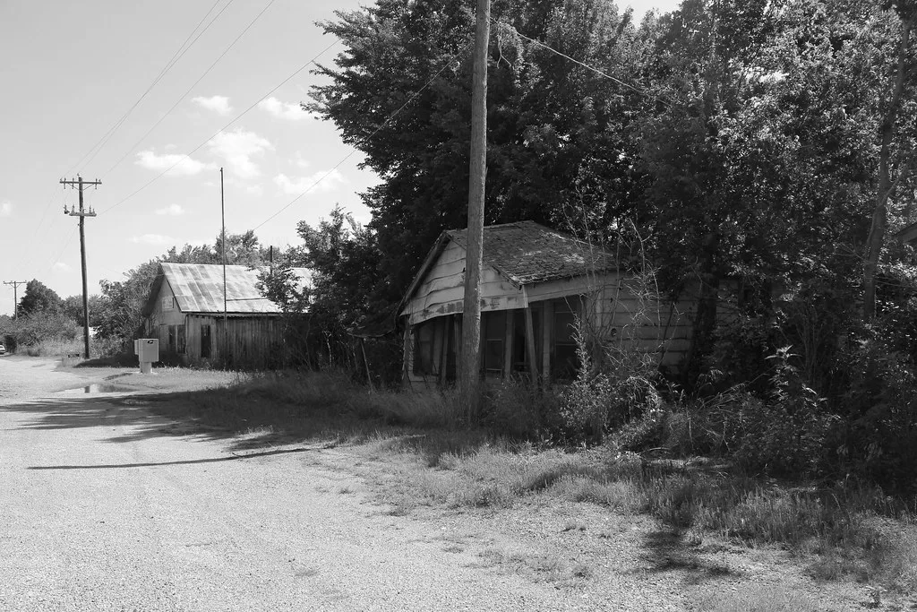 Petite maison abandonnée de bois penchée sous les arbres au bord d’une route, en noir et blanc.