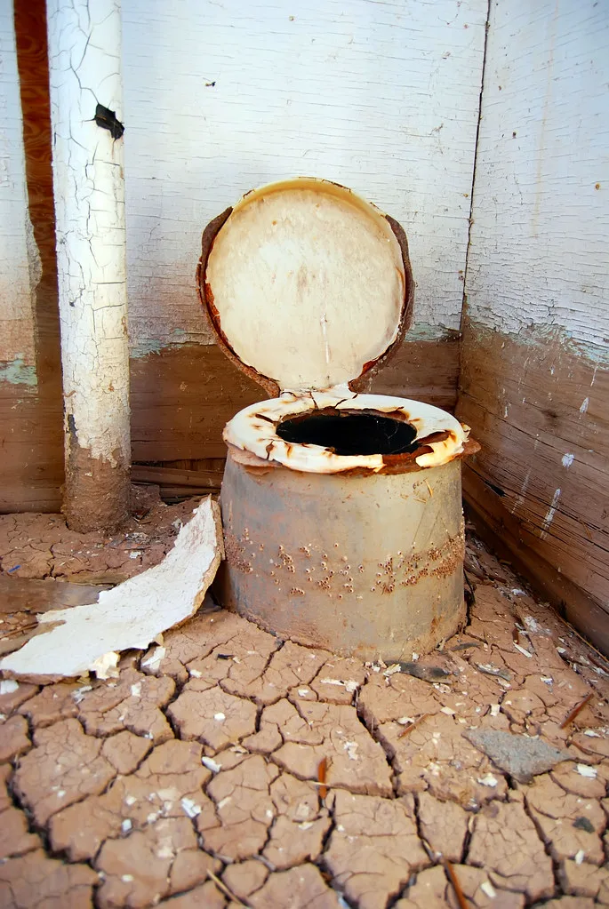 Rusty cylindrical toilet on a cracked floor surrounded by walls with peeling paint.