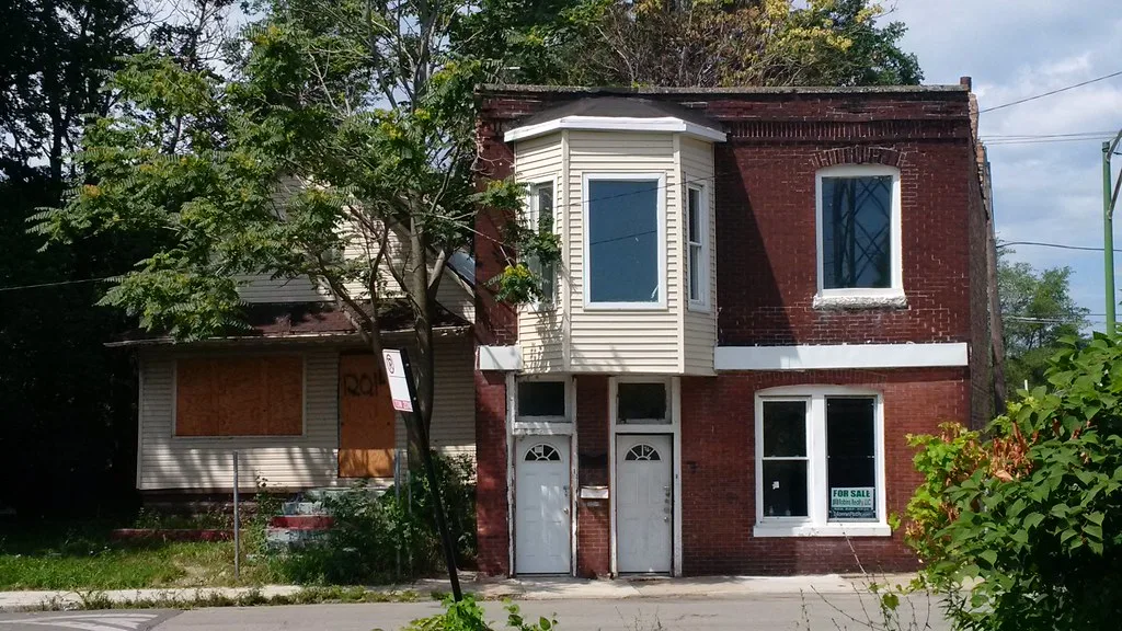 Small house with boarded openings beneath trees beside a red brick house.