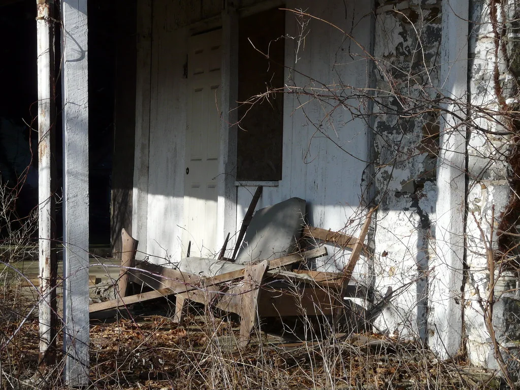 Porche d’une maison délabrée encombré de branches et d’un fauteuil brisé.