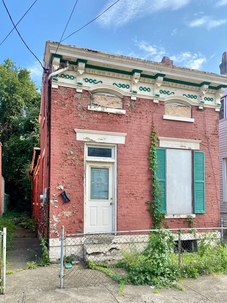 Neglected small pink-brick house with boarded windows and green shutters behind a wire fence.