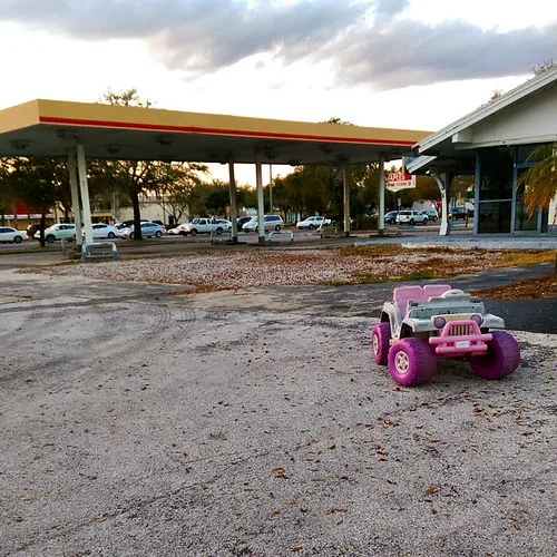 Ancienne station-service sans pompes avec une petite voiture-jouet rose au premier plan.
