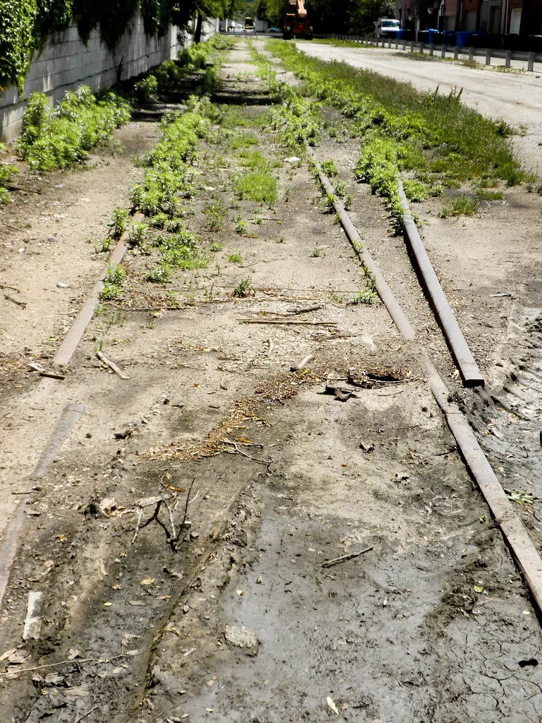 Abandoned railway track with disconnected rails and buried sleepers among weeds beside a street.